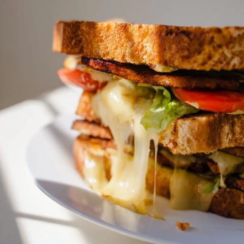 A close-up view of a Vegan Bacon BLT Grilled Cheese cut in half, revealing layers of plant-based ingredients on a rustic wooden cutting board.